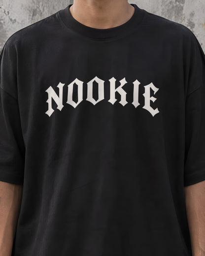 Close do logo NOOKIE em letra gótica branco osso, com 28 cm de largura, no peito da camiseta preta